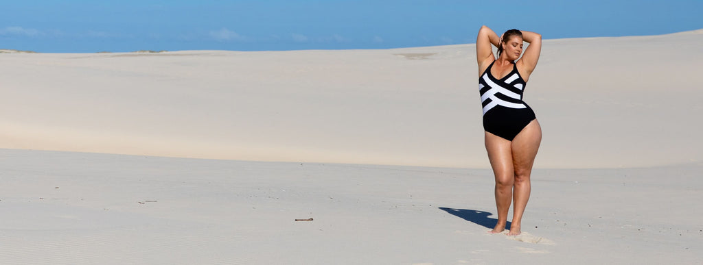 Woman poses on a sand dune wearing a black and white criss cross one piece swimsuit