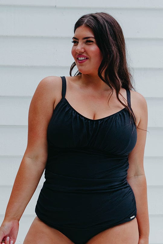 Brunette woman wears a plus size, underwire, black tankini top. It has thin straps, a round neckline, a ruched bustier and tummy area. It fits snug with a luxurious textured fabric.